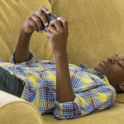 A boy lays on a couch watching something on a hand-held screen device.